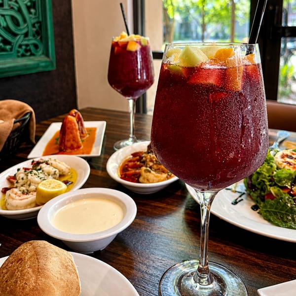 Refreshing red fruit cocktails with citrus garnish surround a restaurant table filled with assorted dishes, sauces, bread, and salad.