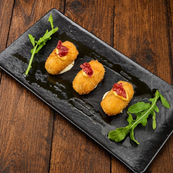 Three golden breaded croquettes topped with creamy sauce and cured meat, garnished with arugula on a black plate.