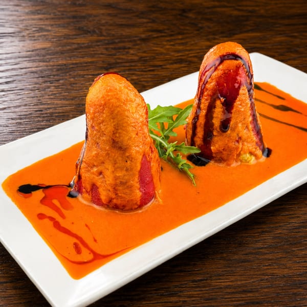 Two golden-brown stuffed croquettes garnished with arugula, drizzled with dark sauce, and served over bright orange sauce.