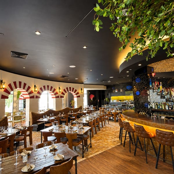 Elegant restaurant interior with set dining tables, arched windows, decorative greenery, and a glowing curved bar.