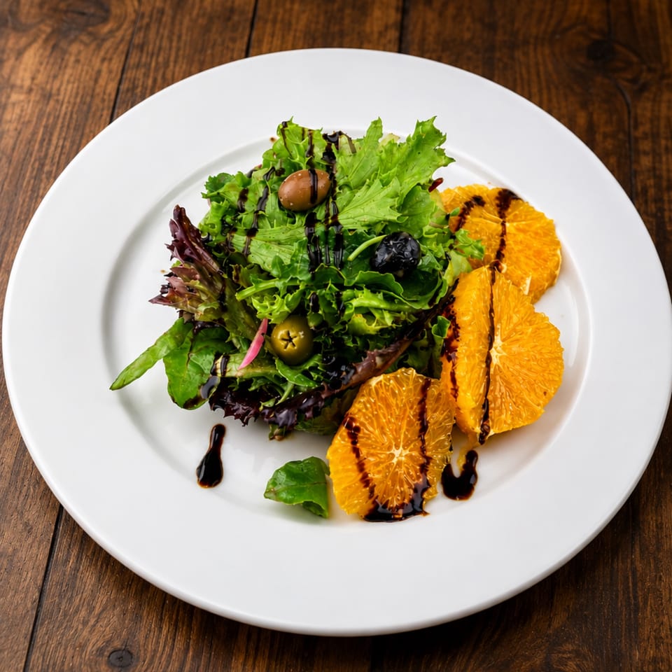Mixed green salad with olives and orange slices drizzled with balsamic glaze on a white plate.