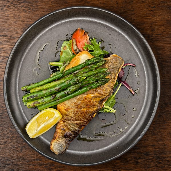 Grilled fish fillet with asparagus, greens, roasted vegetables, and lemon wedge on a dark plate.