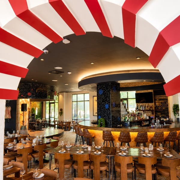 Upscale restaurant interior with red-and-white ceiling accents, set dining tables, curved illuminated bar, and leather seating.