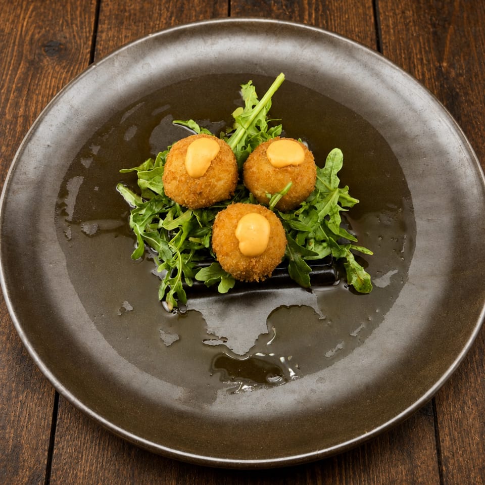 Three golden croquettes topped with creamy sauce, served over fresh arugula on a dark plate with drizzled oil.