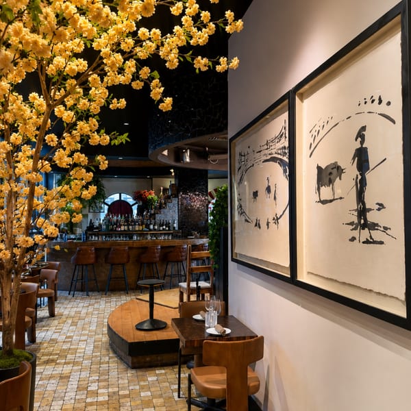 Elegant restaurant interior with flower-filled tree, wooden bar, leather seating, tiled floors, and framed pastoral artwork.