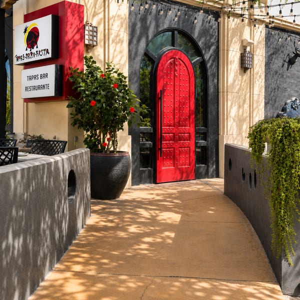 Restaurant entrance with a bright red arched door, tapas bar signage, patio seating, string lights, and flowering plants.