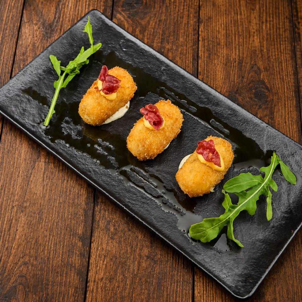 Three golden breaded croquettes topped with cream and cured meat on a black plate with arugula, set on a wooden table.