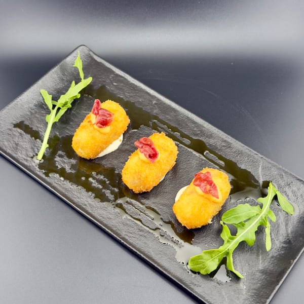 Three golden-fried croquettes with red garnish and sauce, drizzled with oil on a black plate.