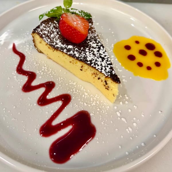 A slice of cheesecake with a strawberry, mint, powdered sugar, and two sauces on a white plate.