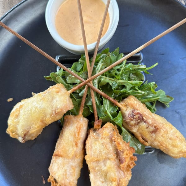 Four fried skewers with a side of arugula and dipping sauce on a black plate.