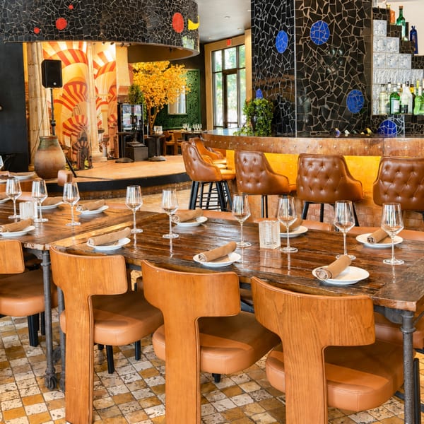 Warmly lit restaurant dining room with set wooden tables, leather chairs, mosaic walls, a bar, and a small stage.