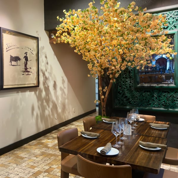 A restaurant interior with a large faux tree, set tables, a bullfighting painting, and an ornate green mirror.