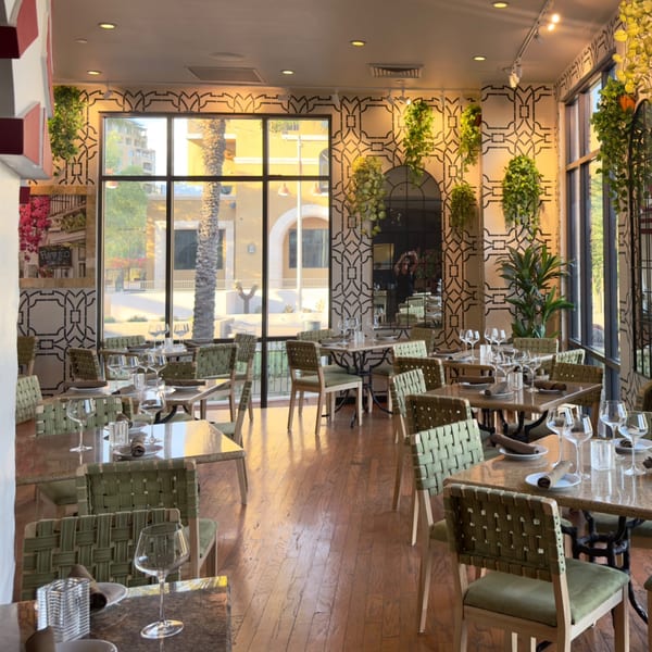 An empty restaurant dining room with set tables, green chairs, and large windows overlooking a sunny street.