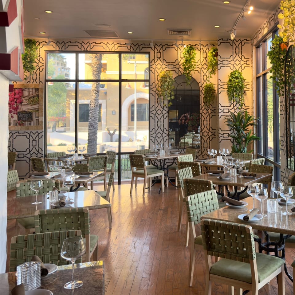An empty restaurant dining room with set tables, green chairs, and large windows overlooking a sunny street.
