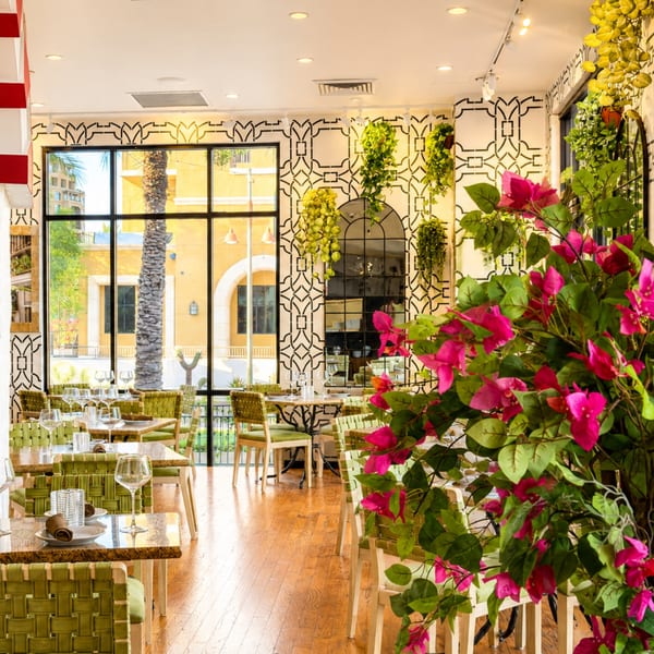 Bright restaurant dining room with green chairs, set tables, patterned walls, large windows, and vibrant pink flowering plants.