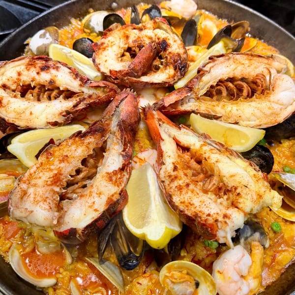 Close-up of a seafood paella with lobster tails, mussels, clams, shrimp, and lemon wedges.