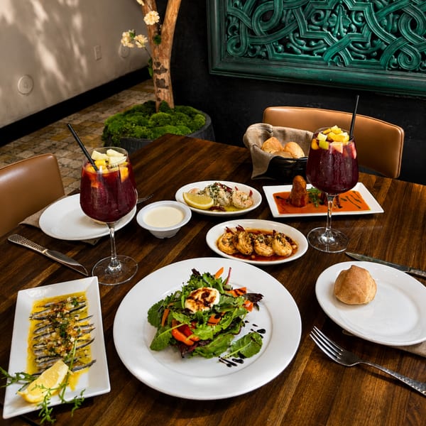 Restaurant table featuring seafood dishes, salad, bread, dipping sauce, and two fruit-topped red cocktails in a warmly decorated dining room.