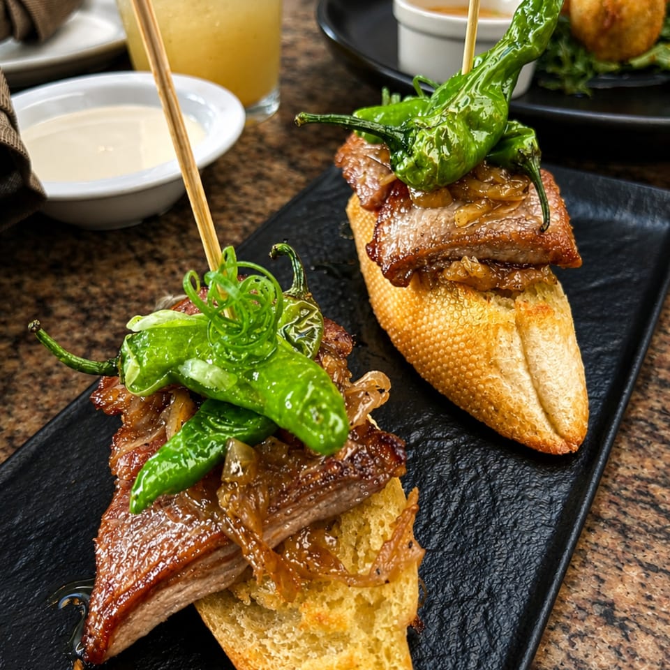 Glazed pork belly and green peppers stacked on toasted baguette slices, served on a black platter with dipping sauce.