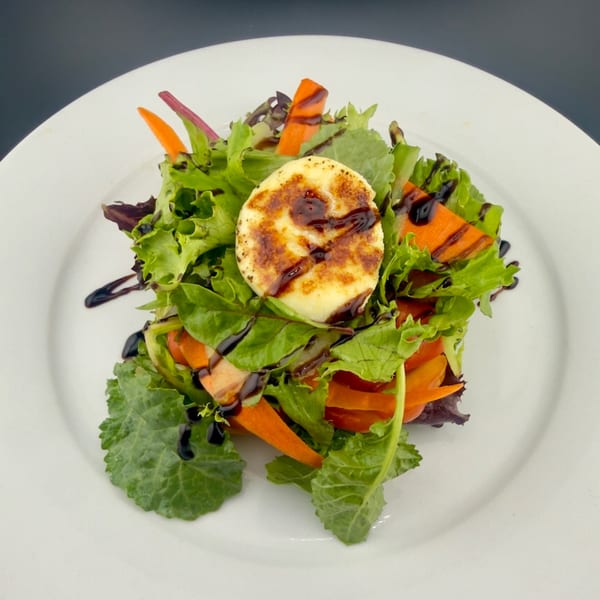 Mixed green salad with grilled halloumi cheese, carrots, and balsamic glaze on a white plate.