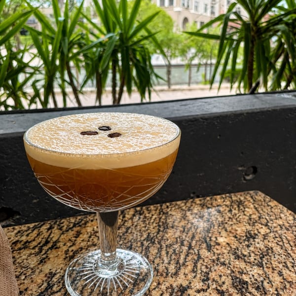 Amber cocktail with foamy topping and coffee beans served in a coupe glass on a granite table overlooking greenery.